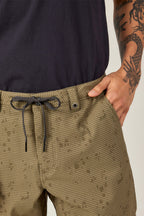 Wearing light olive-green, patterned 686 Everywhere® Hybrid Shorts with a drawstring and black button, paired with a black shirt, a person rests their right hand in the pocket, showing a dragon tattoo on their forearm.