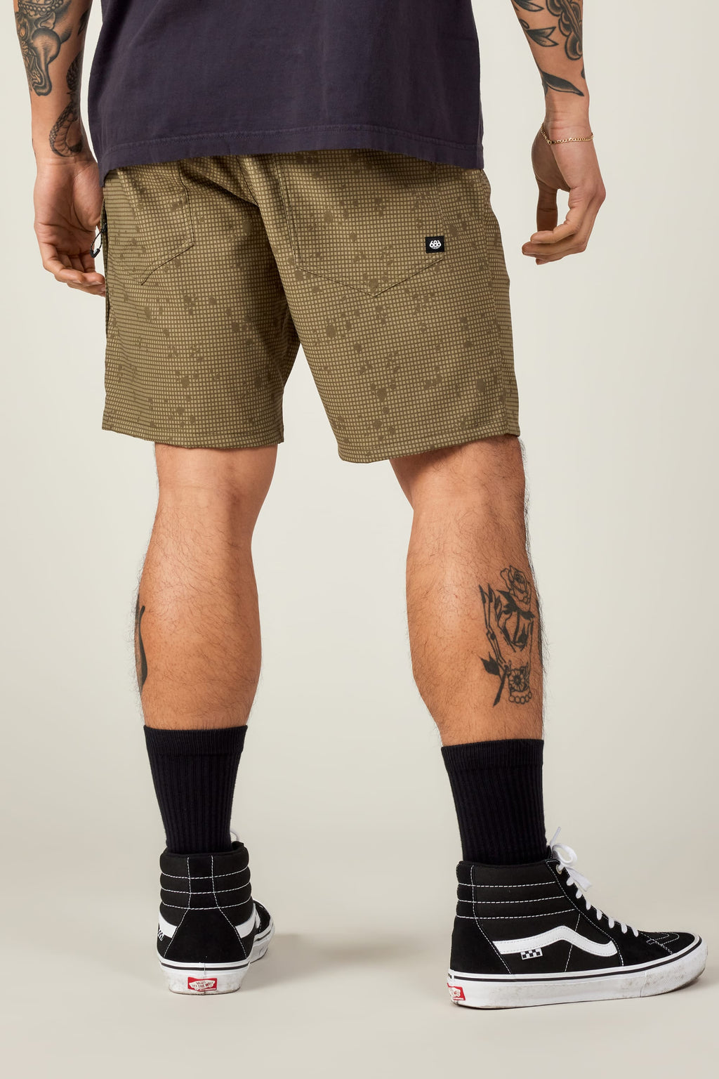 A person seen from behind wears 686 Everywhere® Hybrid Shorts by 686 in olive green checkered, paired with black socks and black high-top Vans. Tattoos cover their arms and legs, including a rose on the calf.