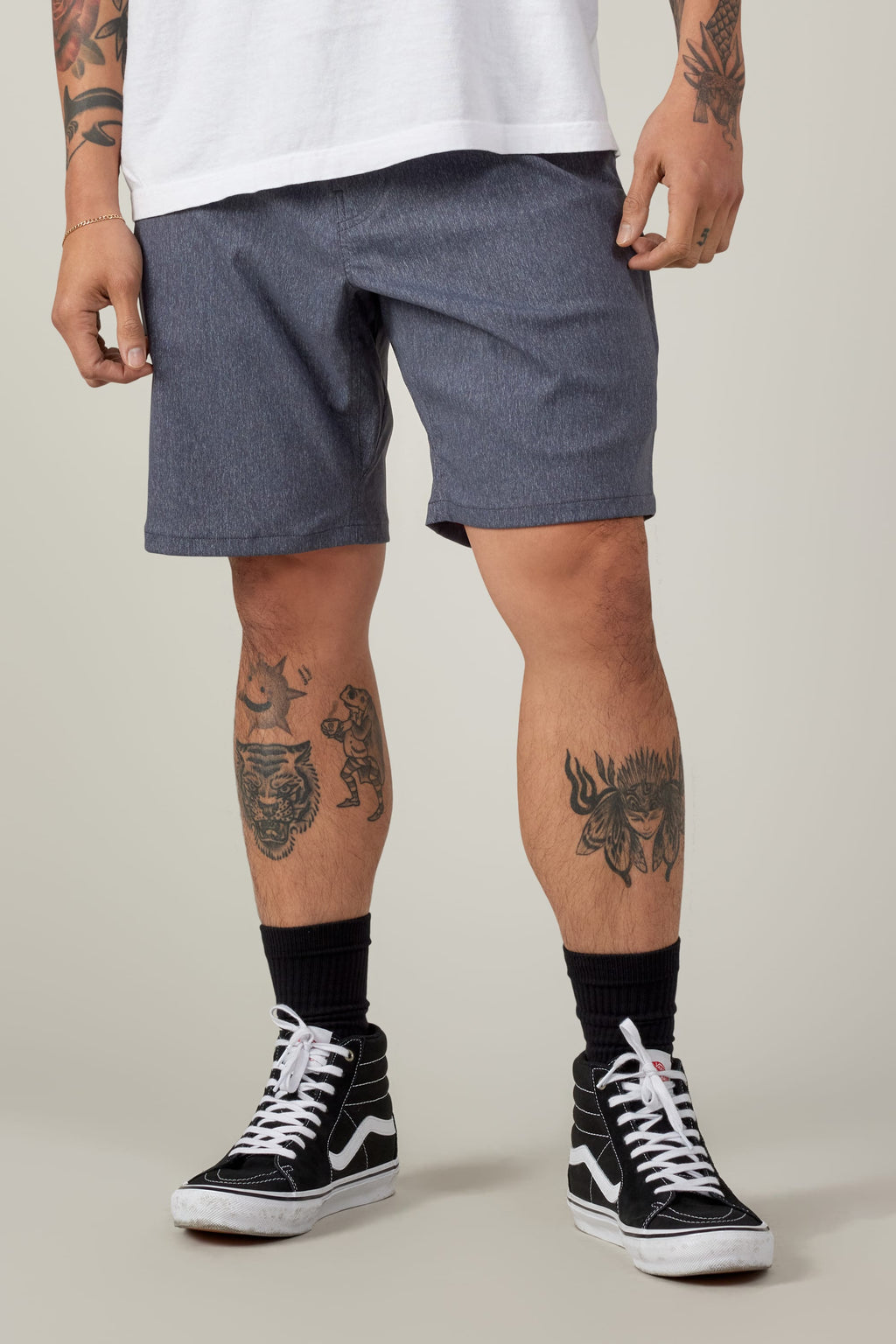 A person wears a white t-shirt, 686 Everywhere® Hybrid Short in blue-gray by 686, black socks, and black high-top sneakers. Tattoos show on their arms and legs. The background is plain and light-colored.
