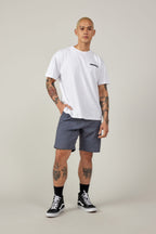 A tattooed person wears a white short-sleeve t-shirt and 686 Everywhere® Hybrid Short by 686, paired with black socks and black high-top sneakers, standing against a plain light background with one hand in their pocket.