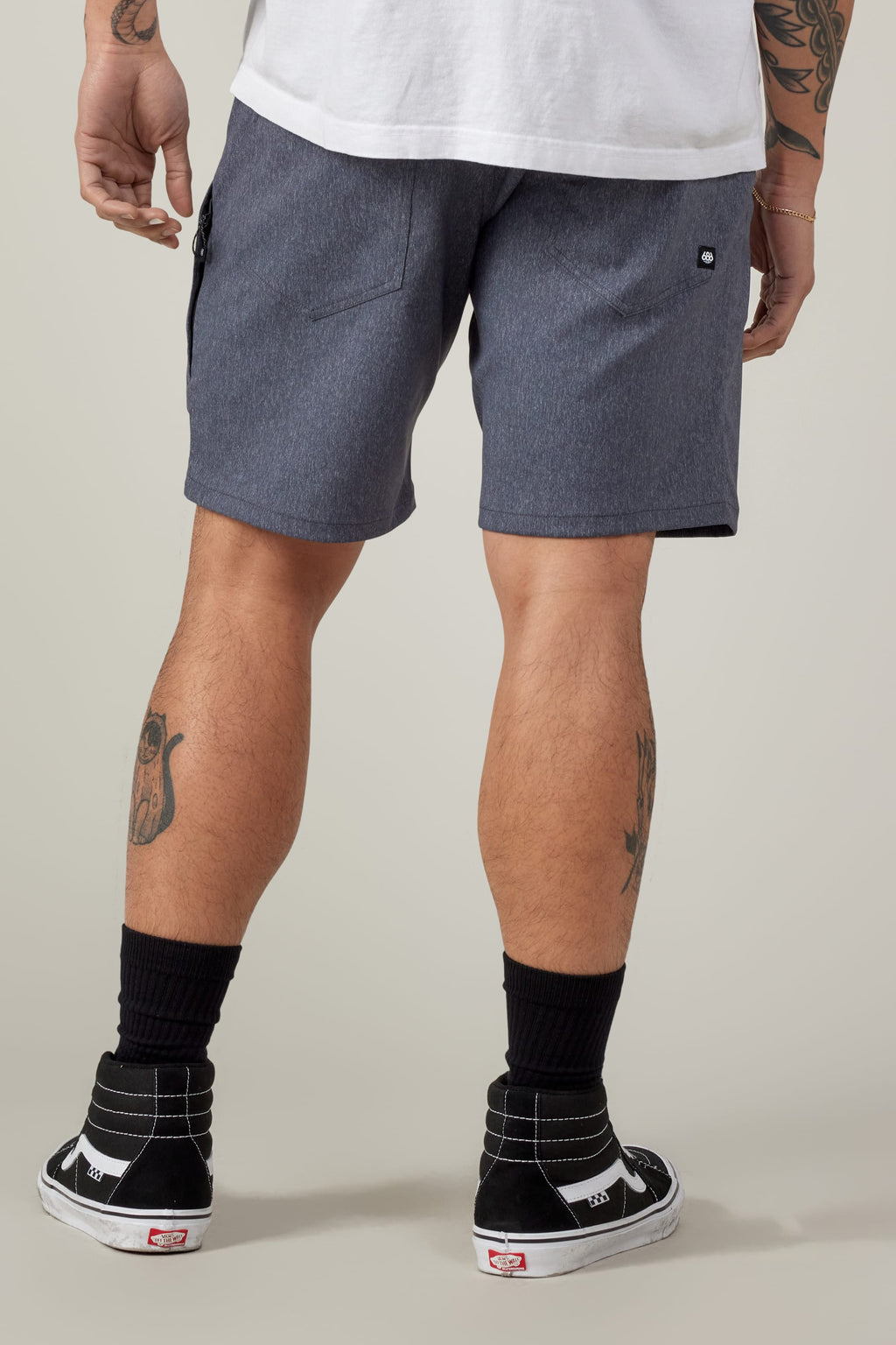 A person in a white T-shirt, 686 Everywhere® Hybrid Short by 686, black socks, and black Vans sneakers stands with their back to the camera, showing leg tattoos.