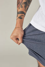 A person with a tattooed arm and gold bracelet pulls on the fabric of their blue 686 Everywhere® Hybrid Short by 686, paired with a white t-shirt, against a plain light background.