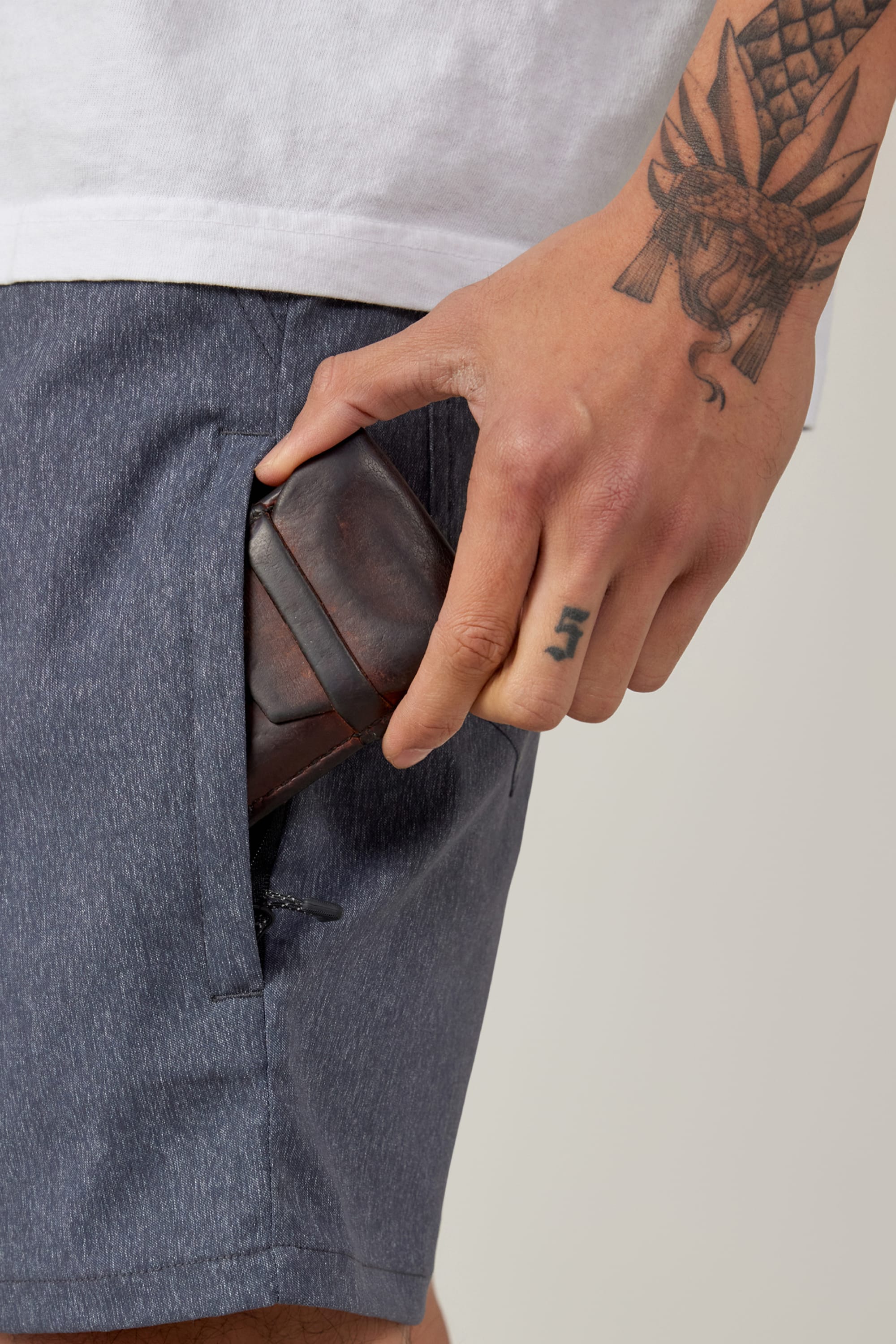 A tattooed individual places a dark brown wallet into the pocket of 686 Everywhere® Hybrid Shorts by 686, styled with a white shirt.