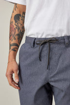 A person wears a white T-shirt and 686 Everywhere® Hybrid Short in blue-gray—breathable and stain-resistant by 686. Their left arm shows a rose and two faces tattoo plus a thin bracelet. Only the lower torso and arm are shown.