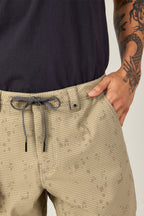 A person wears 686 Men's Everywhere® Hybrid Short in light tan with a pattern, drawstring, and one hand in the pocket, paired with a black shirt. A dragon tattoo is visible on their arm.
