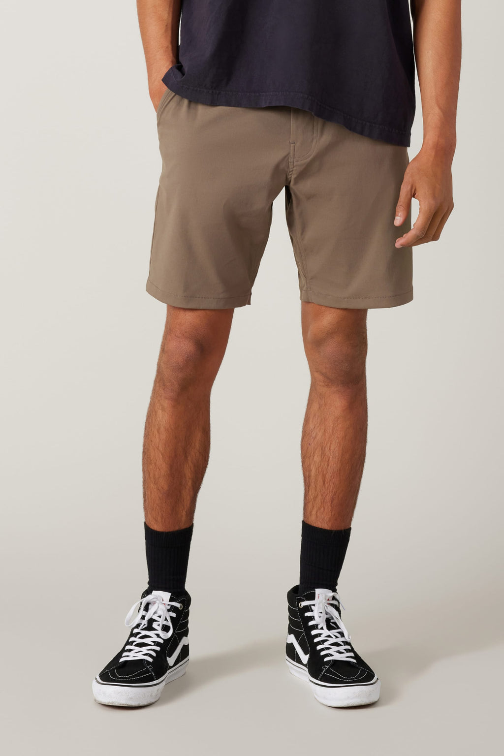 A person wears a dark t-shirt, 686 Everywhere® Hybrid Short in light brown by 686, black crew socks, and black high-top sneakers with white laces, standing against a plain background.