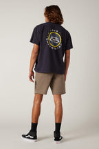 A person with shoulder-length hair stands facing away, wearing a black graphic T-shirt, the 686 Everywhere® Hybrid Short from 686, black socks, and black sneakers with white soles, against a plain light background.