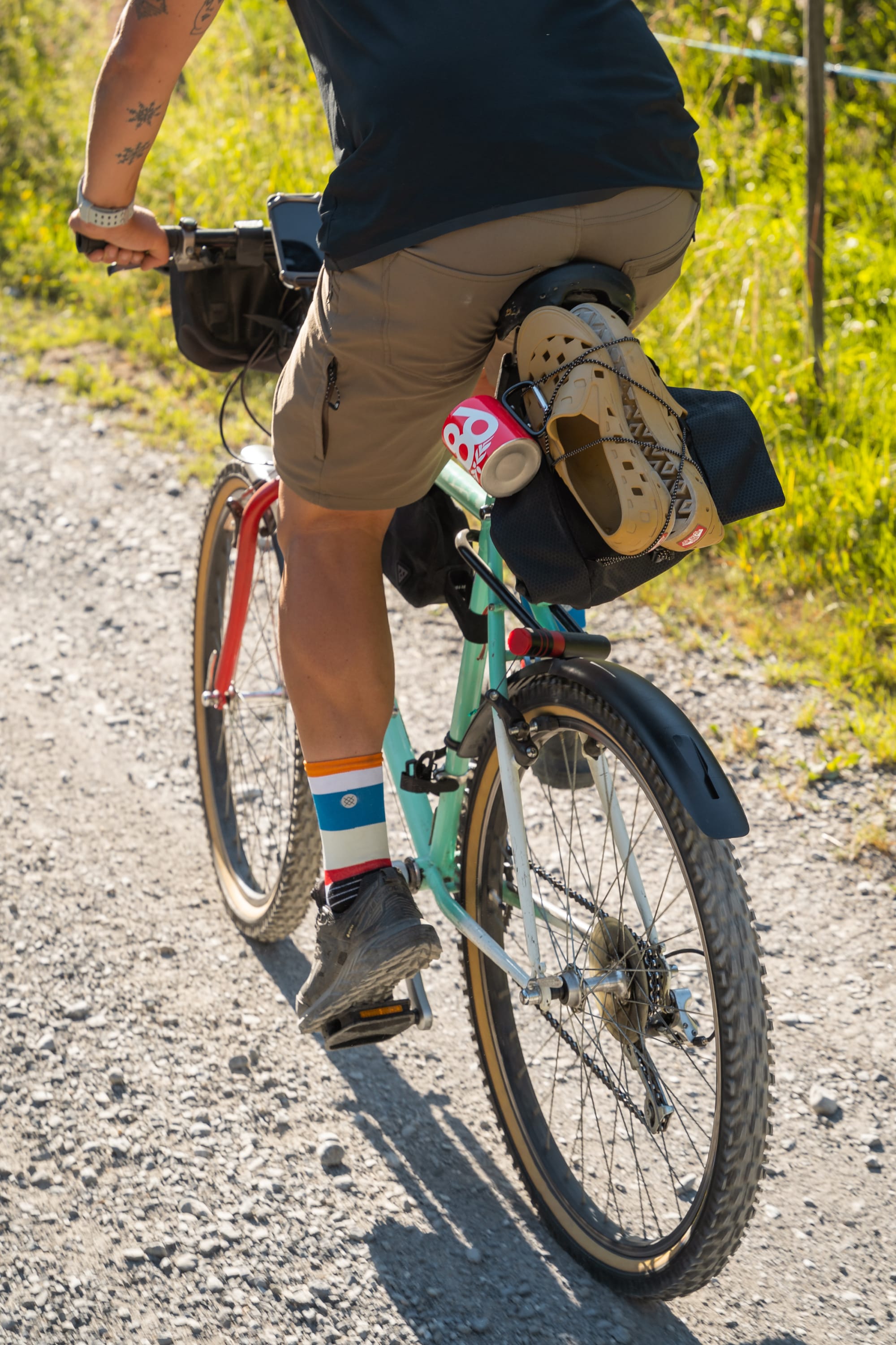 Wearing 686 Everywhere® Hybrid Short by 686 and colorful socks, a cyclist rides a gravel path bordered by green grass, with essentials like a helmet and sleeping pad secured to their bike.
