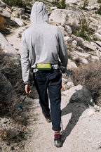 Wearing the 686 Men's Anything Cargo Pant - Slim Fit, a hiker tackles a rocky trail with trekking poles and a small green bag, ready for adventure among shrubs and large rocks.