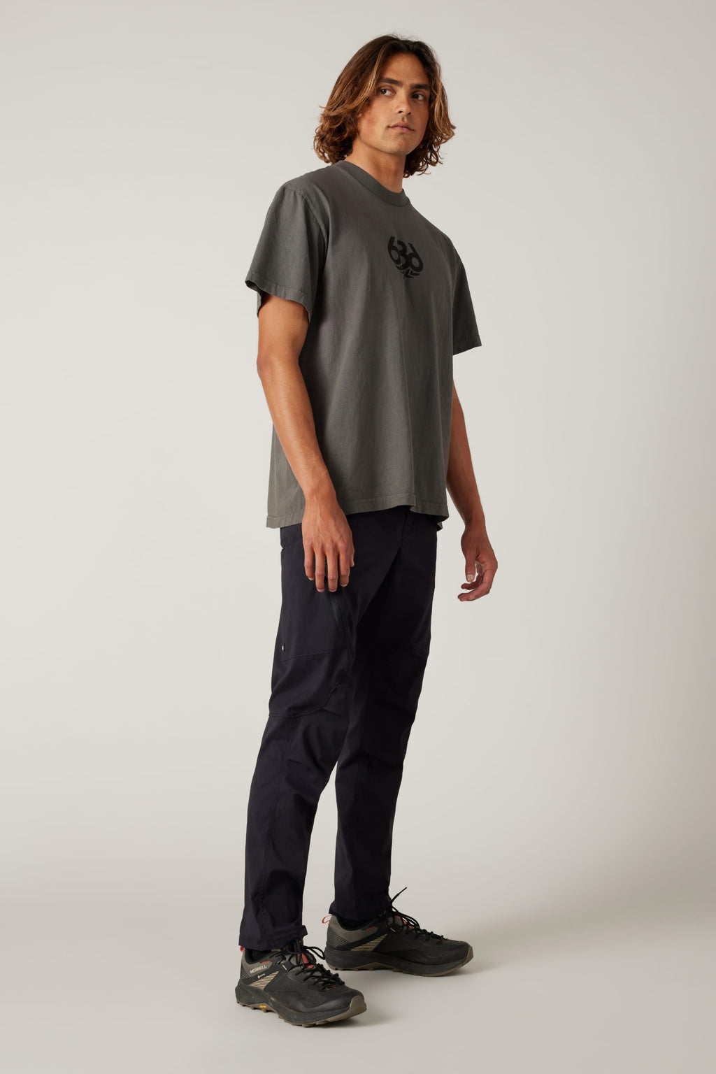 A young man with shoulder-length hair stands at an angle, wearing a loose grey t-shirt, 686 Men's Anything Cargo Pant - Slim Fit by 686, and black sneakers against a plain light background.