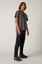 A young man with shoulder-length hair stands at an angle, wearing a loose grey t-shirt, 686 Men's Anything Cargo Pant - Slim Fit by 686, and black sneakers against a plain light background.
