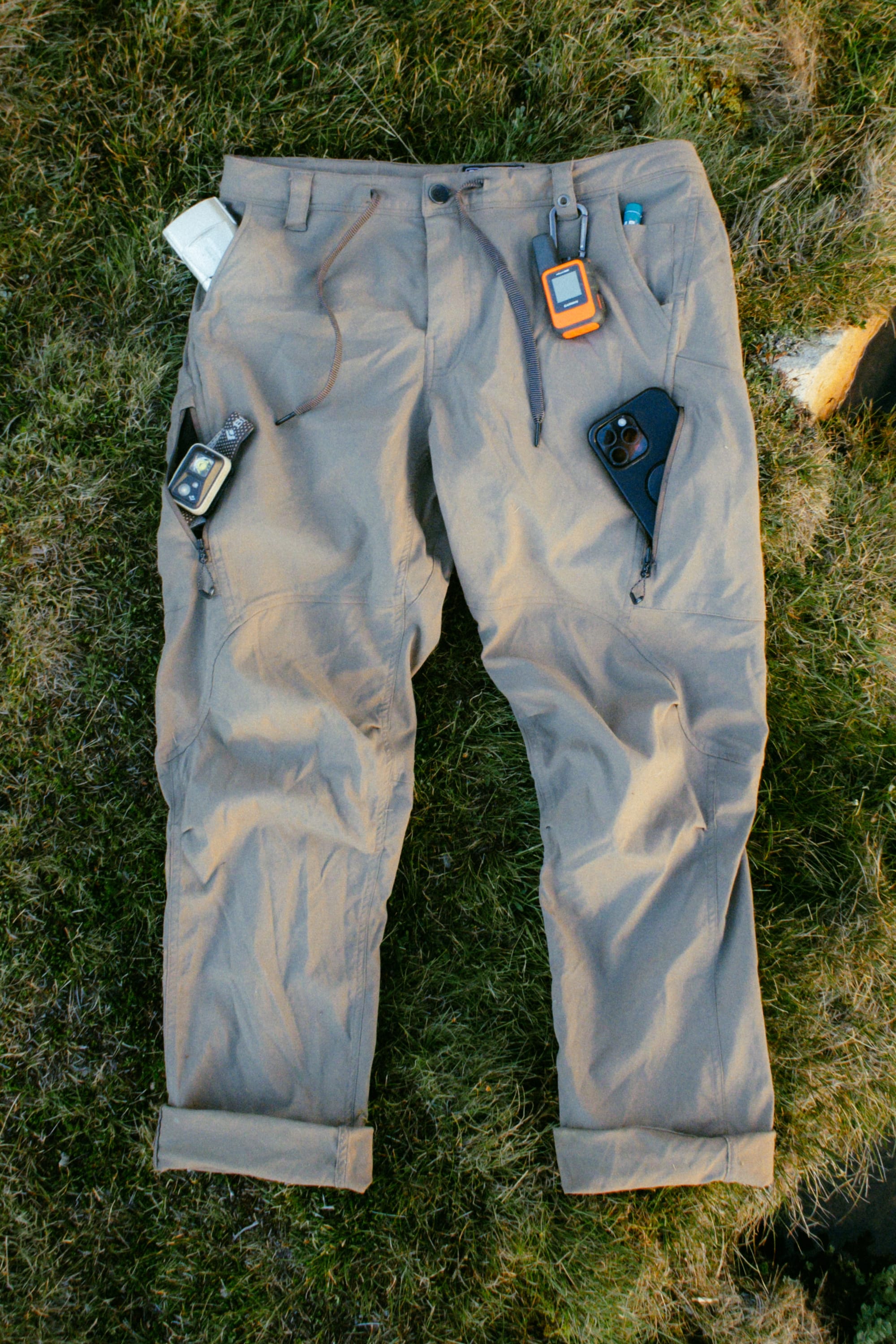 The 686 Men's Anything Cargo Pant - Slim Fit in light brown rests on grass, with adventure gadgets—such as a smartphone, GPS device, and satellite communicator—stored in its 13 pockets and attached to the belt loops.