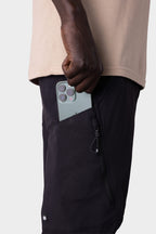 A man in a beige shirt and black 686 Men's Anything Cargo Pant - Slim Fit slips a triple-camera smartphone into the zippered side pocket, ready for any adventure. The background is plain light gray.