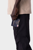 A person in a beige shirt and 686 Men's Anything Cargo Pant - Slim Fit slides a passport into one of its 13 pockets. The shot highlights their hand, the pocket, and passport against a plain background, capturing an adventurous vibe.