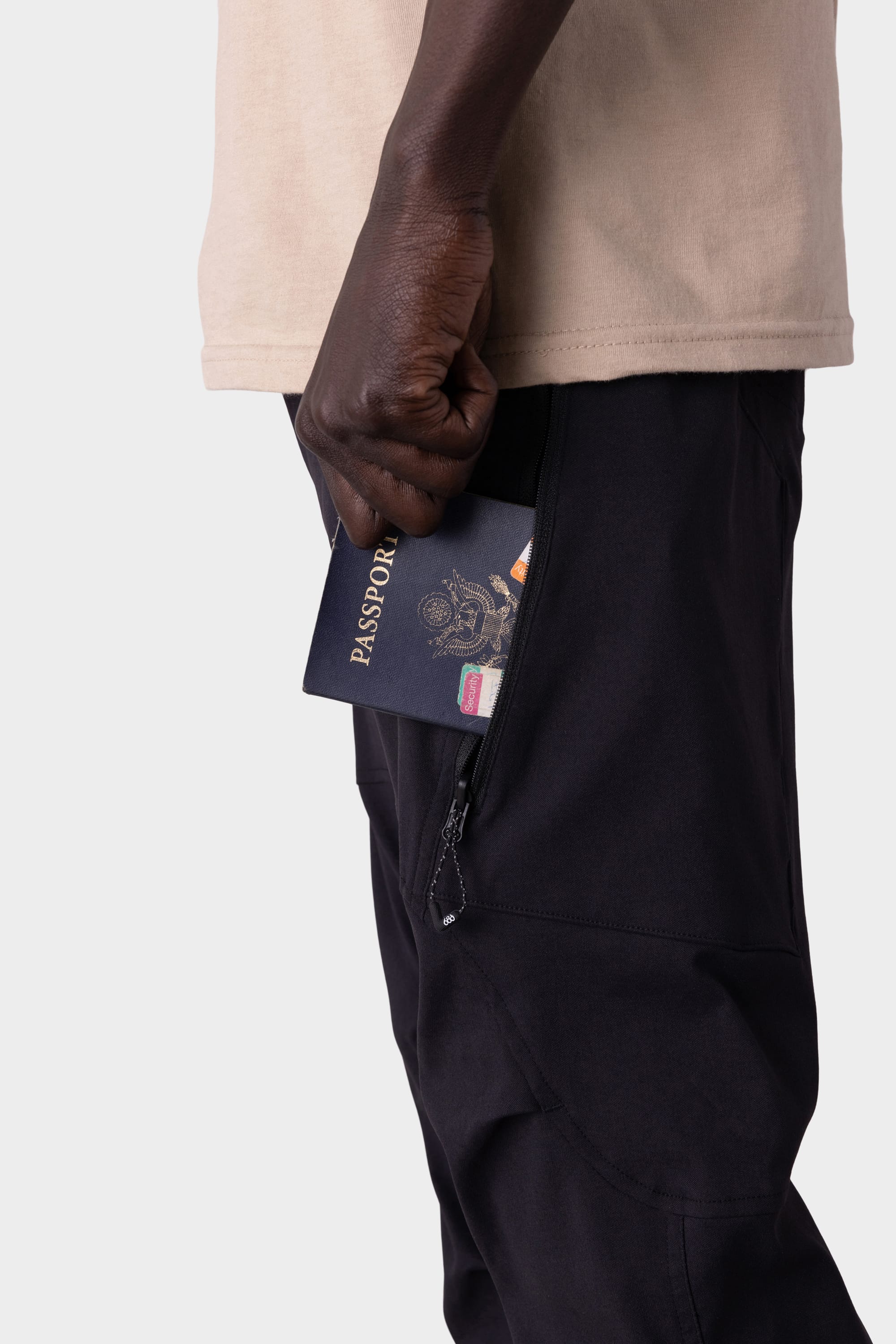 A person in a beige shirt and 686 Men's Anything Cargo Pant - Slim Fit slides a passport into one of its 13 pockets. The shot highlights their hand, the pocket, and passport against a plain background, capturing an adventurous vibe.