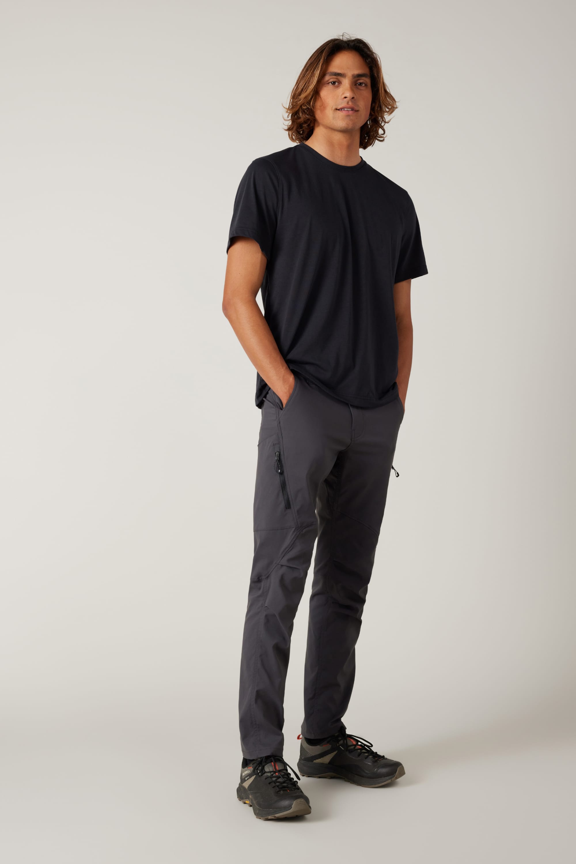 A person with shoulder-length brown hair stands with hands in pockets, wearing a black t-shirt, 686 Men's Anything Cargo Pant - Slim Fit by 686, and black sneakers against a plain light background, ready for any adventure.