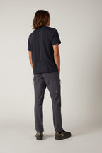 A person with shoulder-length hair stands facing away in a black t-shirt, 686 Men's Anything Cargo Pant - Slim Fit by 686, and black shoes against a plain light background—ready for any adventure.