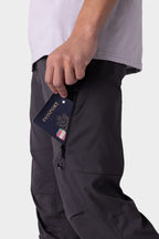 Wearing the 686 Men's Anything Cargo Pant - Slim Fit, someone in dark pants and a light shirt slips a passport into the thigh zipper pocket, ready for their next adventure. The background is plain and light-colored.