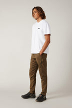 A person with long brown hair wears a white T-shirt, 686 Men's Anything Cargo Pant - Slim Fit by 686, and black sneakers, standing with hands in pockets against a plain light background.
