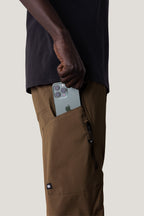 Wearing 686 Men's Anything Cargo Pant - Slim Fit by 686 and a black shirt, someone slides a green triple-camera smartphone into a zippered side pocket, preparing for their next adventure.