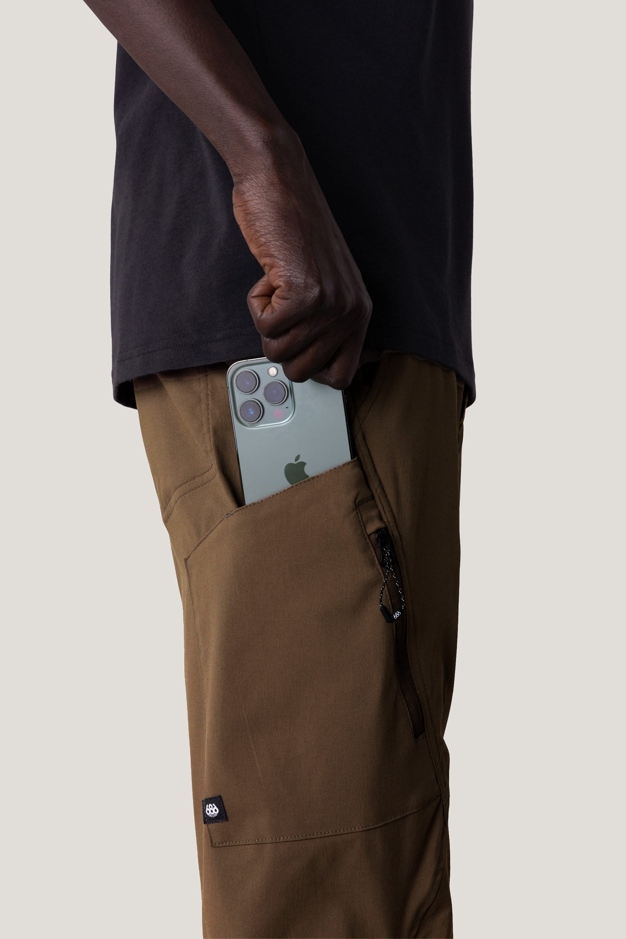 Wearing 686 Men's Anything Cargo Pant - Slim Fit by 686 and a black shirt, someone slides a green triple-camera smartphone into a zippered side pocket, preparing for their next adventure.