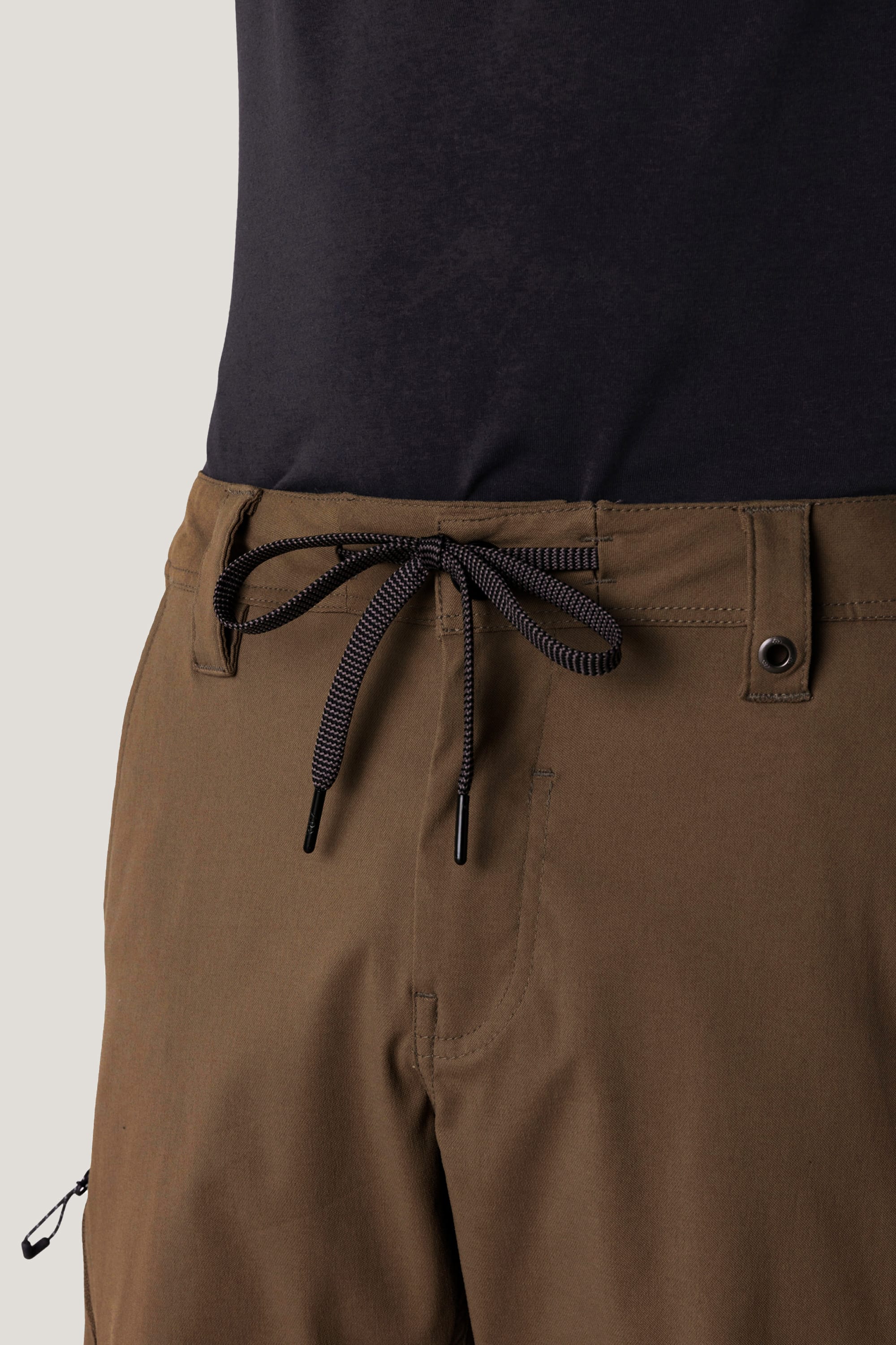 Close-up of a person wearing 686 Men's Anything Cargo Pant - Slim Fit in brown with a black drawstring, single waist button, and black shirt. The 13-pocket design by 686 is visible. Photo cropped at torso and upper thighs.
