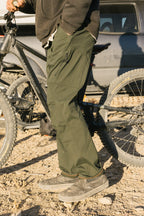 Wearing 686 Men's Anything Cargo Pant - Slim Fit in olive green and gray slip-on shoes, a person stands by a black mountain bike on dirt, with a car in the background. The scene is outdoors under sunny skies, evoking adventure.