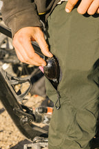 A person slips sunglasses into the zippered pocket of their 686 Men's Anything Cargo Pant - Slim Fit, with a bicycle in the background—ready for adventure in these technical cargo pants by 686.