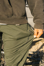 Wearing 686 Men's Anything Cargo Pant - Slim Fit in green, a person stands by a bike, hand on the thigh pocket zipper of their 13-pocket pants, ready for adventure.
