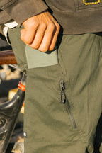 A person slips a green wallet into the zippered pocket of the 686 Men's Anything Cargo Pant - Slim Fit, with part of a bike and rocky ground in the background, hinting at adventure.