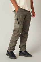 Wearing the 686 Men's Anything Cargo Pant - Slim Fit by 686, featuring a 13-pocket design and zippered side pocket, and a cream tee with black sneakers, a person stands with one hand in their pocket against a neutral background.