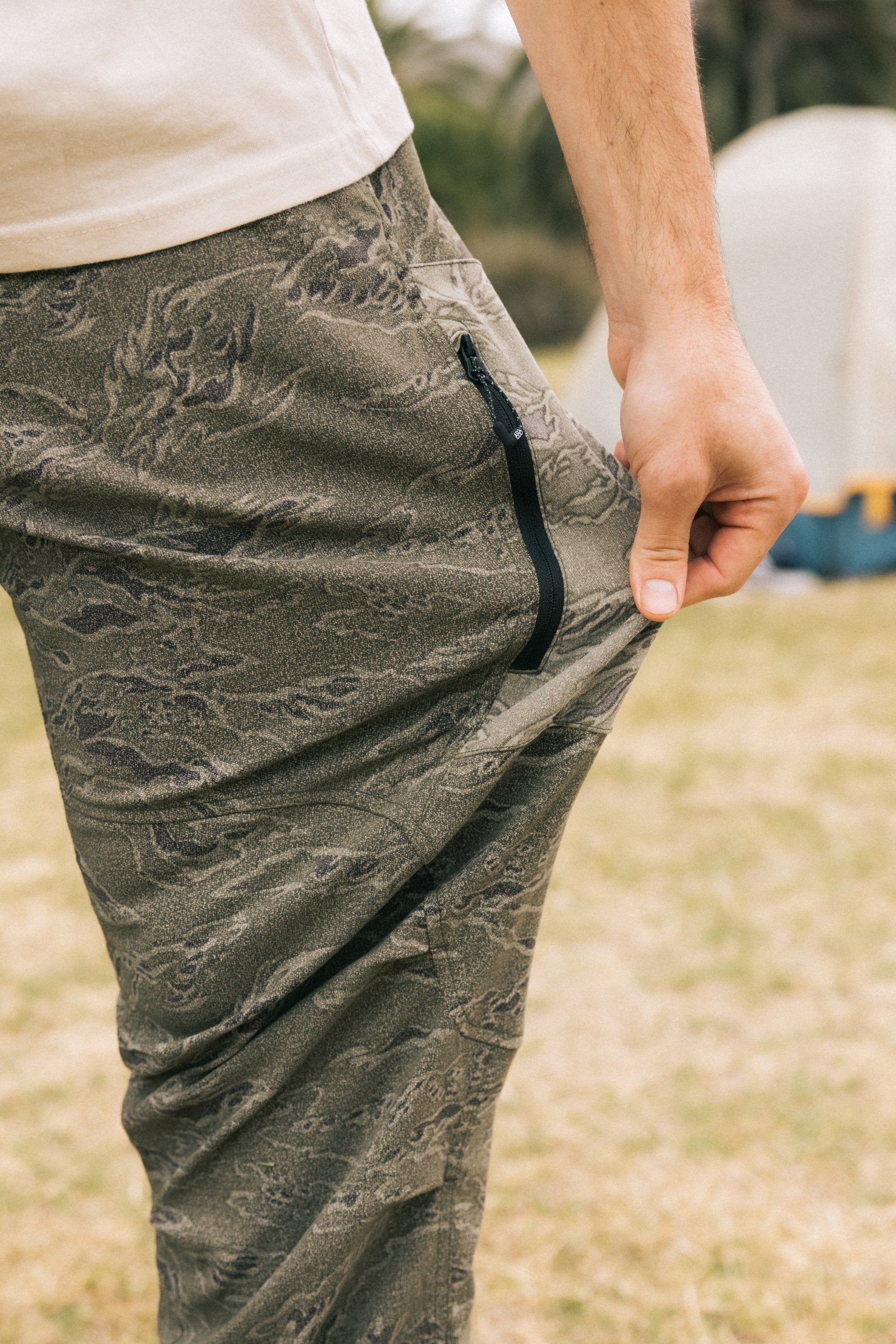 A person wears the 686 Men's Anything Cargo Pant - Slim Fit in olive green, showing off its stretch and black zipper pocket. Outdoors on grass with a tent blurred behind, these 686 pants are built for any adventure.