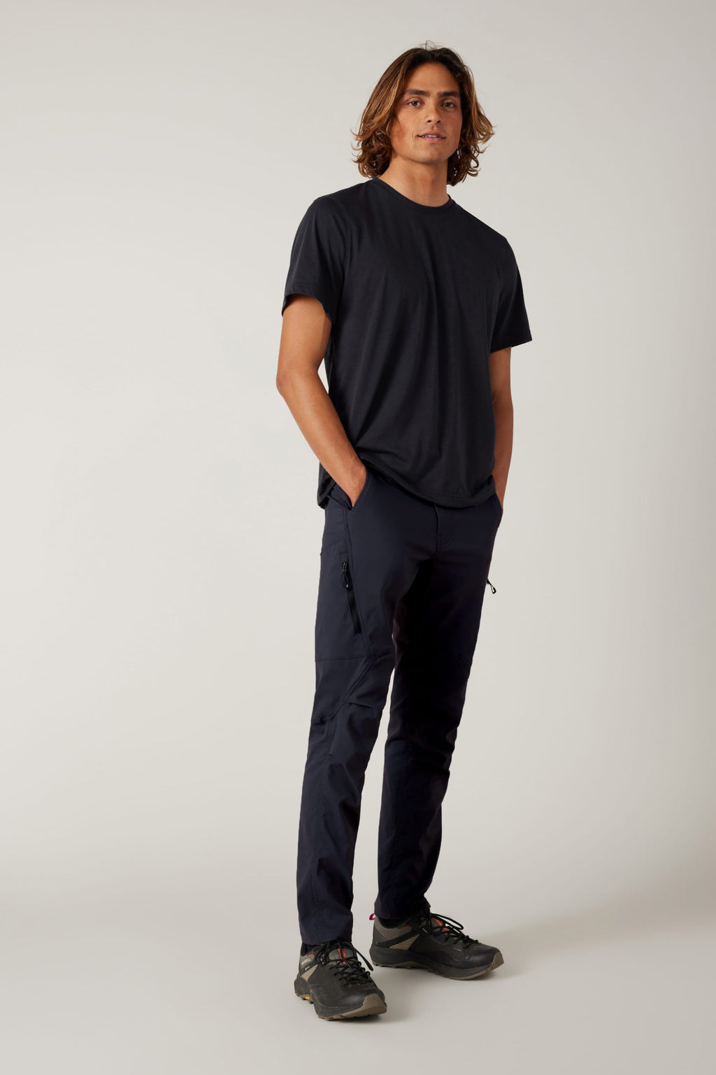 A young man with wavy brown hair stands confidently against a plain background, wearing a black t-shirt, black sneakers, and the 686 Men's Anything Cargo Pant - Slim Fit by 686. His hands are in his pockets, ready for any adventure.