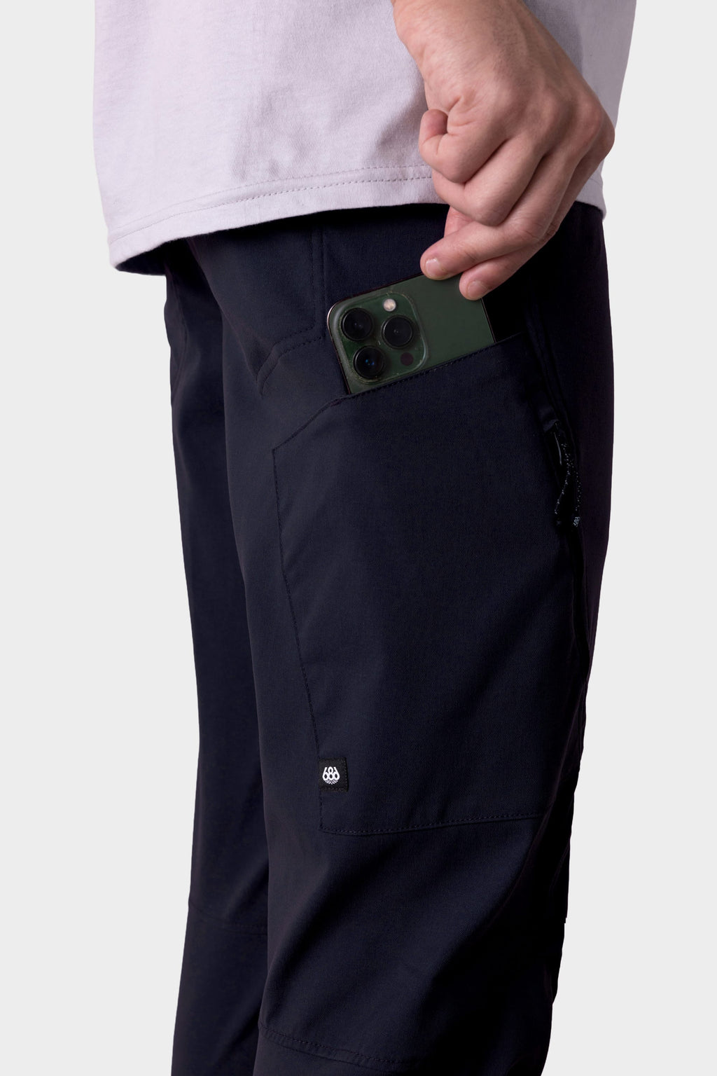 A person puts a green smartphone into the zippered side pocket of the 686 Men's Anything Cargo Pant - Slim Fit by 686, ready for their next adventure.