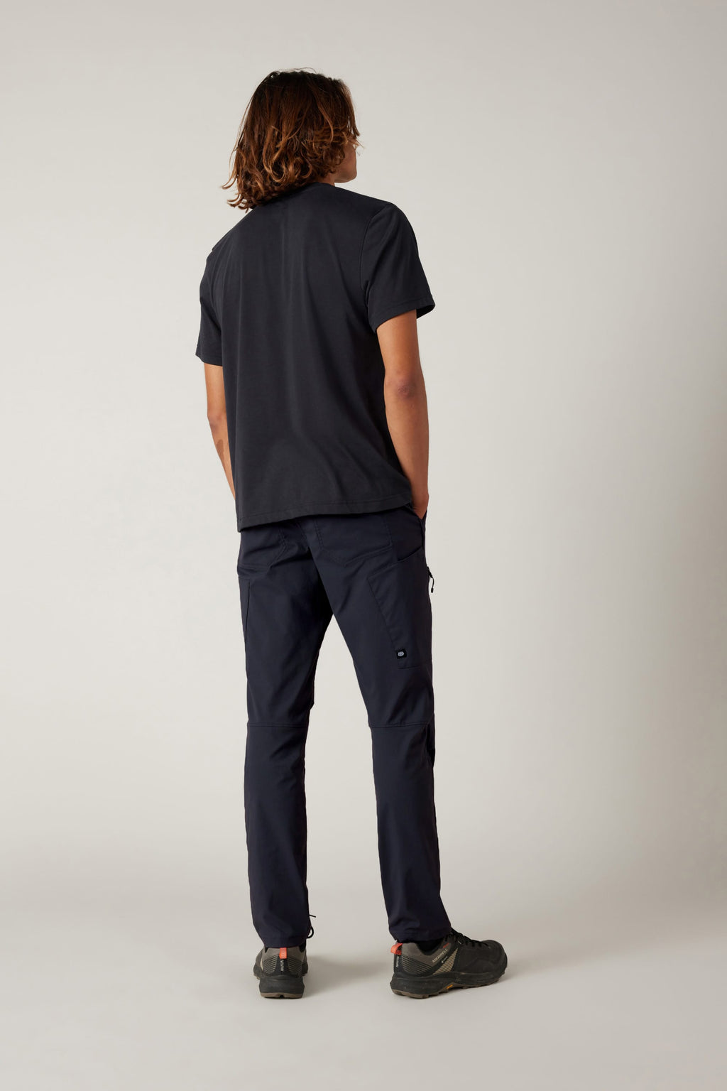 A person with shoulder-length hair faces away in a black short-sleeve shirt, 686 Men's Anything Cargo Pant - Slim Fit by 686, and black shoes—ready for any adventure against a plain light background.