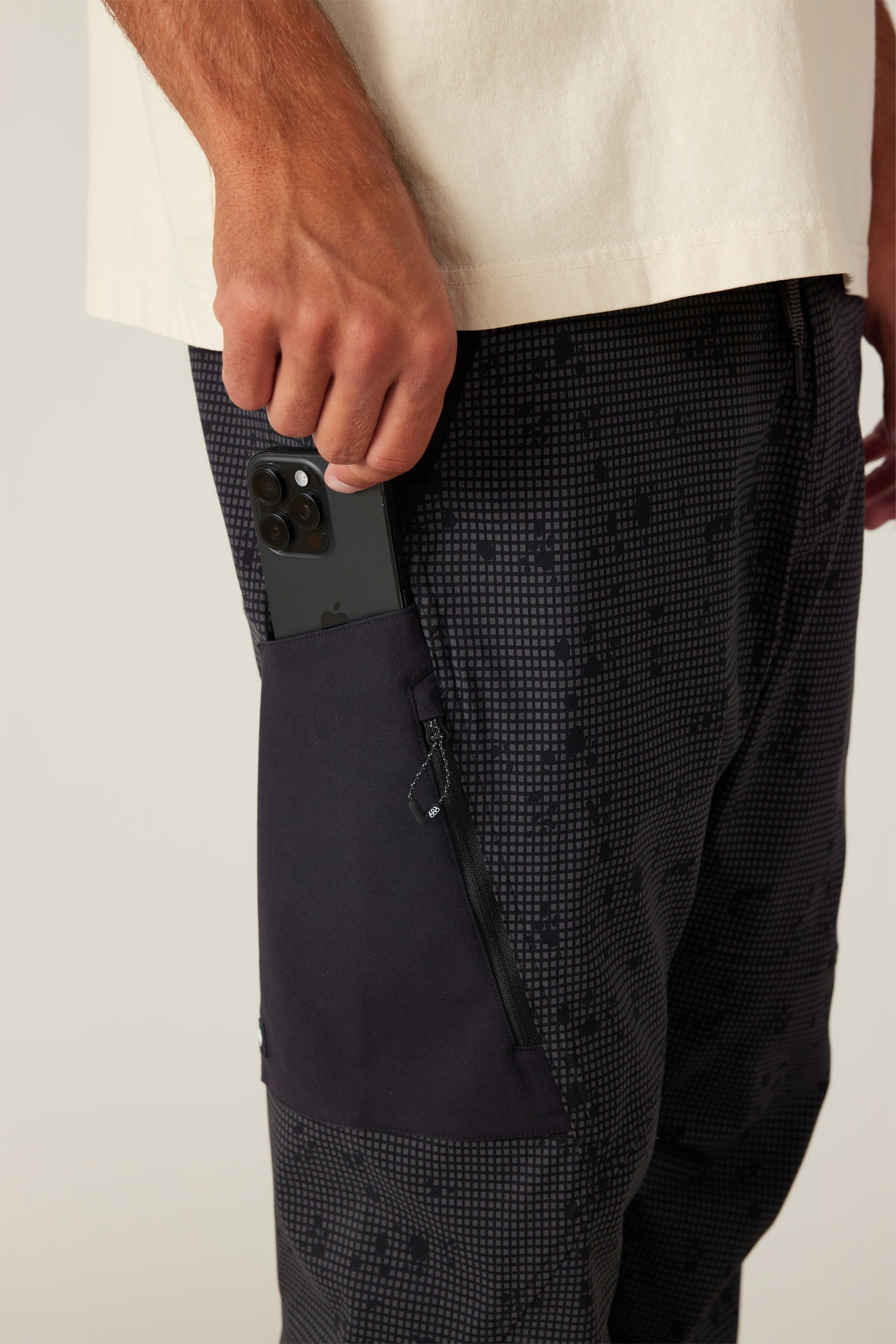 Wearing 686 Men's Anything Cargo Pant - Slim Fit, a person slips a black smartphone into the zippered side pocket. Only their hand, hip, and upper legs are visible, showcasing 686's readiness for any adventure.
