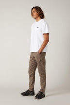 A person with long wavy hair stands against a plain background, wearing a white T-shirt and 686 Men's Anything Cargo Pant - Slim Fit by 686, paired with black sneakers. Hands in pockets, they look off to the side, ready for adventure.