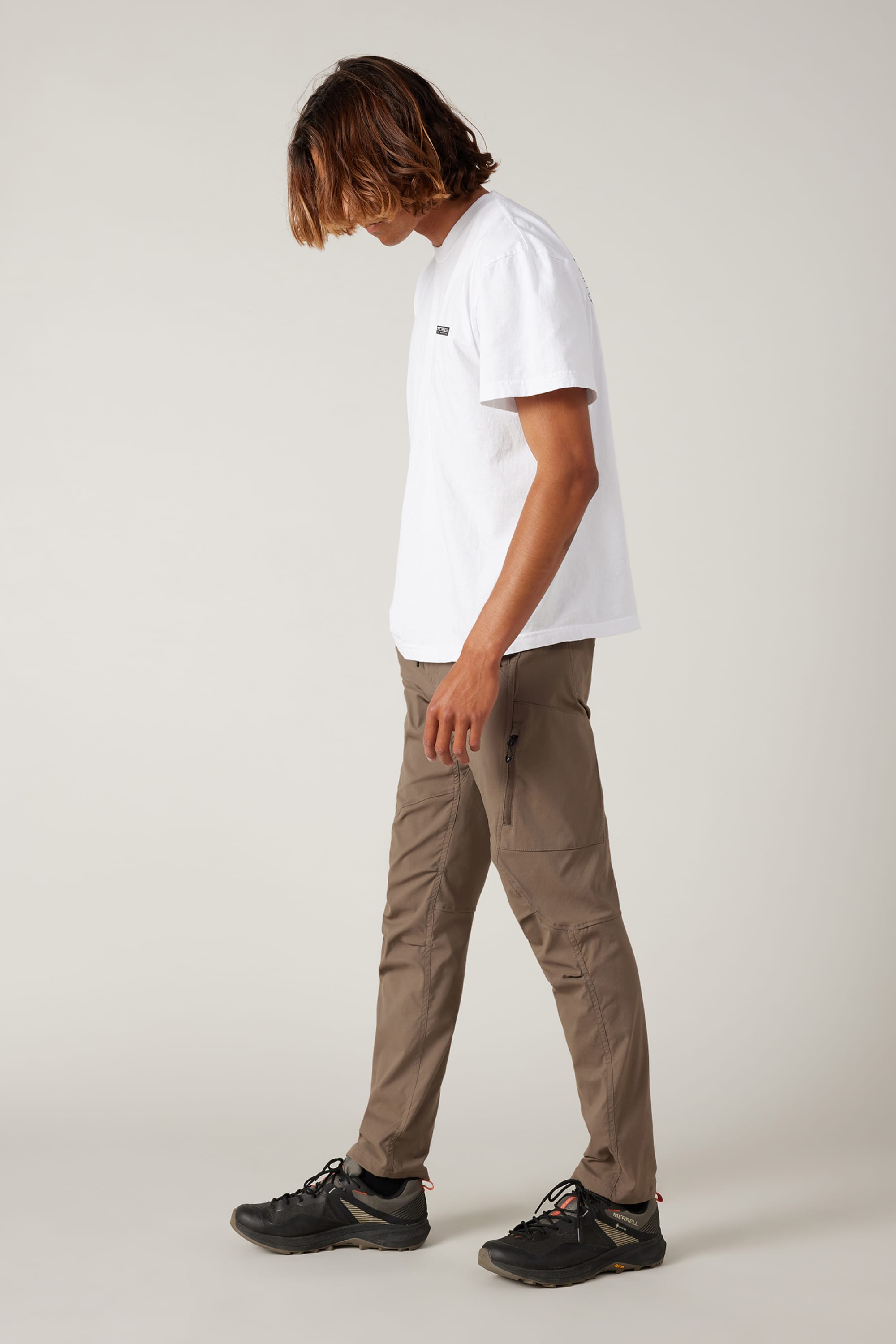 A person with shoulder-length hair in a white t-shirt, 686 Men's Anything Cargo Pant - Slim Fit by 686, and black sneakers stands against a plain light gray background, looking down with one arm relaxed, ready for their next adventure.