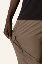 A man in 686 Men's Anything Cargo Pant - Slim Fit (tan) and a black shirt stretches the side to reveal a zippered pocket—one of 13 total—making these 686 pants ideal for any adventure.