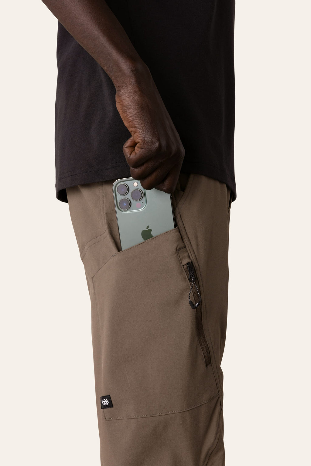 Wearing the 686 Men's Anything Cargo Pant - Slim Fit by 686, someone slips a green triple-camera iPhone into a zippered pocket. The 13-pocket design is ideal for adventure, and a black shirt completes their look.