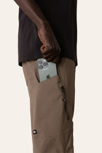 Wearing the 686 Men's Anything Cargo Pant - Slim Fit by 686, someone slips a green triple-camera iPhone into a zippered pocket. The 13-pocket design is ideal for adventure, and a black shirt completes their look.
