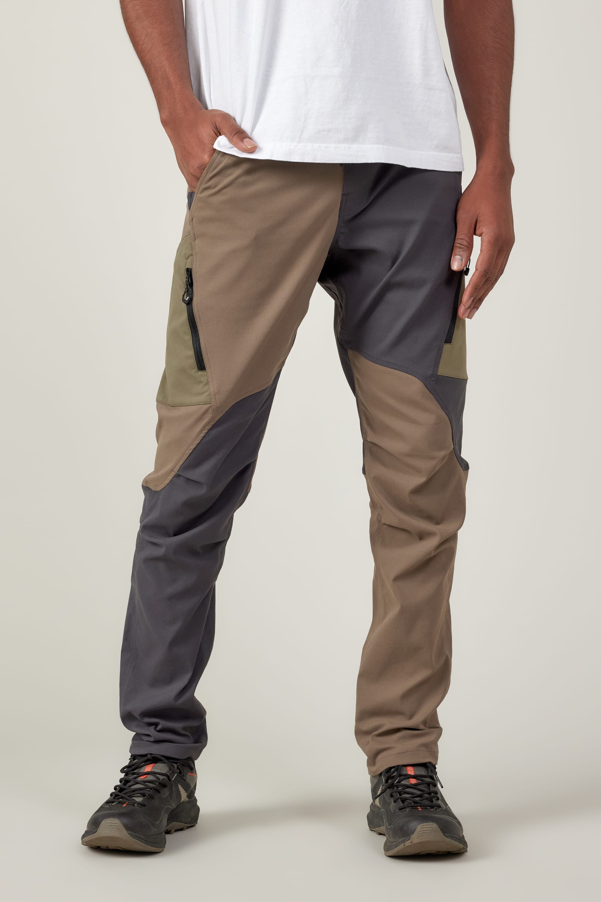 Wearing a white t-shirt and 686 Men's Anything Cargo Pant - Slim Fit by 686 in gray, brown, and olive, paired with black hiking shoes, a person stands with one hand in their pocket against a plain background, ready for adventure.