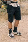 A person wearing a beige shirt, black 686 Men's Everywhere® Hybrid Short - Relaxed Fit, black Vans socks, and black-and-white Vans sneakers stands outdoors on a dirt path among dry grass and cactus plants.