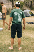 A man stands barefoot on grass, facing away, wearing a breathable green graphic tee, a white cap, and 686 Men's Everywhere® Hybrid Short - Relaxed Fit by 686. Behind him are another man, tents, and palm trees at a campsite.