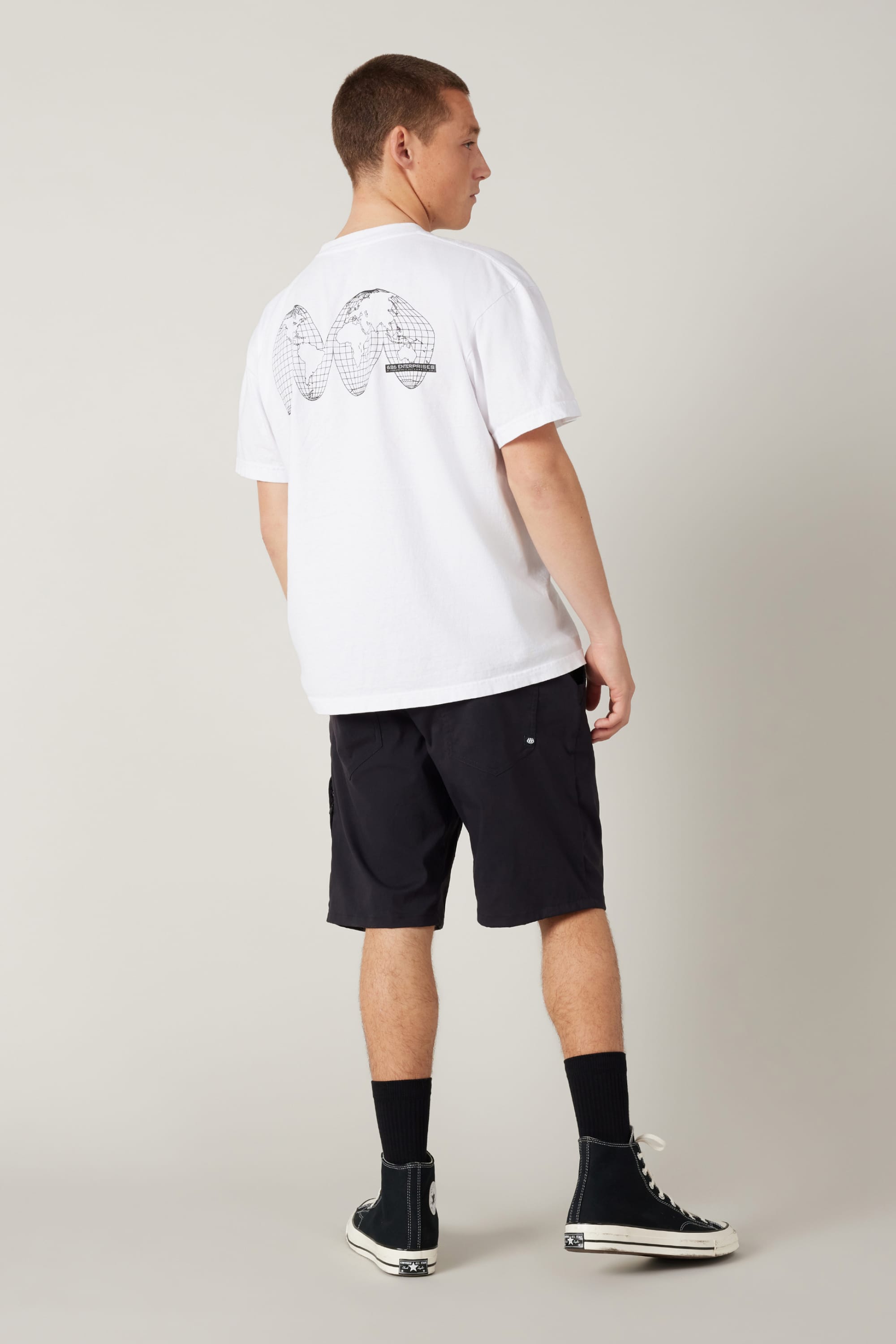 A person faces away, wearing a white tee with black globe and WAV graphic, 686 Men's Everywhere® Hybrid Short - Relaxed Fit, black socks, and black high-top sneakers against a plain background.