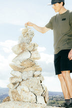 A person in a cap, green t-shirt, and 686 Men's Everywhere® Hybrid Short - Relaxed Fit is adding a stone to the top of a tall rock pile outdoors beneath a bright, partly cloudy sky.