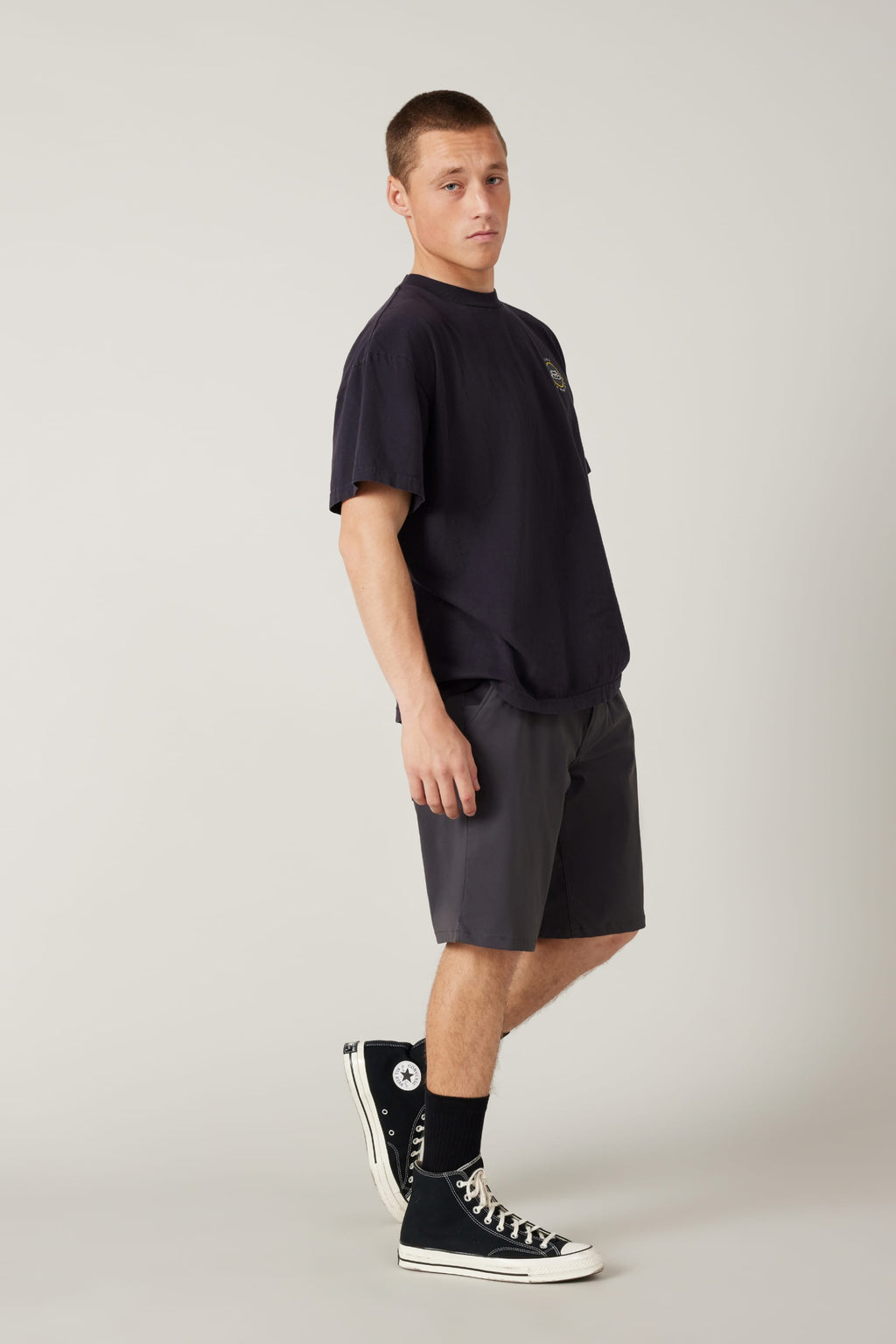 A young man stands against a plain background, wearing a loose black T-shirt, 686 Men's Everywhere® Hybrid Short - Relaxed Fit by 686, black socks, and black high-top Converse sneakers, looking at the camera with a neutral expression.
