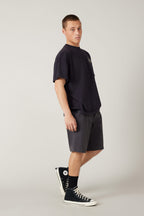 A young man stands against a plain background, wearing a loose black T-shirt, 686 Men's Everywhere® Hybrid Short - Relaxed Fit by 686, black socks, and black high-top Converse sneakers, looking at the camera with a neutral expression.