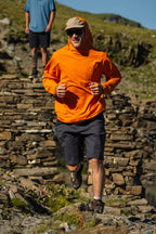 Wearing 686 Men's Everywhere® Hybrid Short - Relaxed Fit, a person jogs outdoors on a rocky path in an orange hoodie, sunglasses, and cap. Another stands behind with hills and stone ruins in the background.