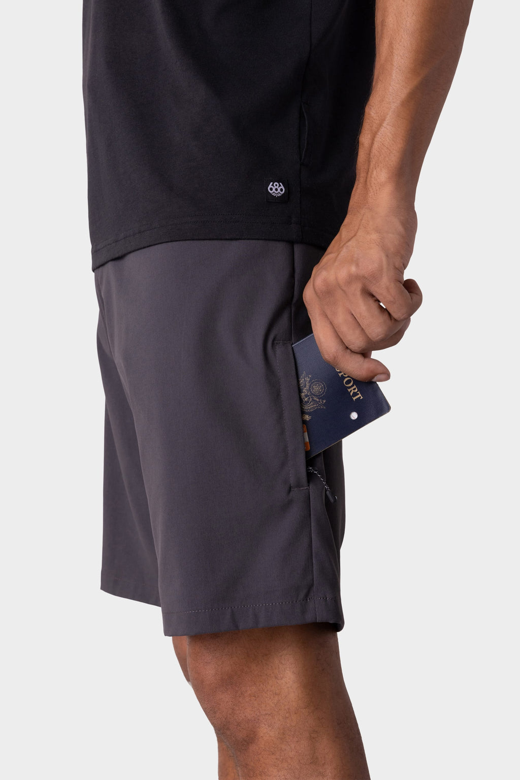A person in a dark t-shirt and 686 Men's Everywhere® Hybrid Short - Relaxed Fit (gray) holds a blue passport partly tucked into the shorts’ zippered side pocket. The background is plain and light-colored.